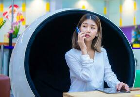 Beautiful asian business woman sitting on modern round chair and using phone to communicate with customer with serious face while looking to the left side, Digital marketing. photo