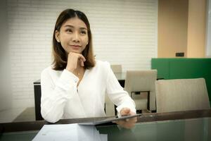 Beautiful asian office girl in close-up shot is sitting pretending to think in copy space with a doubtful expression and holding tablet on her desk. photo