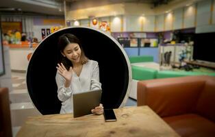 Attractive asian businesswoman greeting customers through tablet while sitting in modern round chair with smile in company. photo