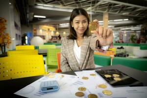 hermosa asiático mujer de negocios es mostrado participación un bitcoin en su mano mientras sonriente con un documento visible en su escritorio en su oficina. foto