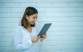 Pretty asian business woman stands sideways and looks at the tablet she is holding with a smile with white bricks in the background, Digital marketing. photo