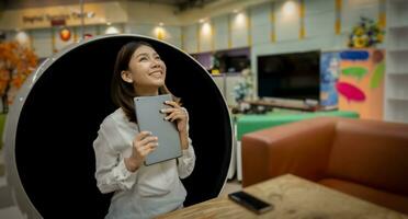 Beautiful asian businesswoman is overjoyed when she gets promoted by her boss via tablet with a smile while sitting on a modern round chair in the living room. photo