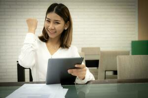 Beautiful asian office girl wearing white dress is sitting showing fist while holding tablet with smile on her face, Business woman cheering concept. photo