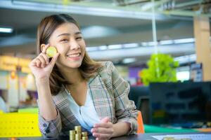 Young beautiful asian business woman holding a golden coin in one hand to her cheek while looking to the camera with smile on her face. photo