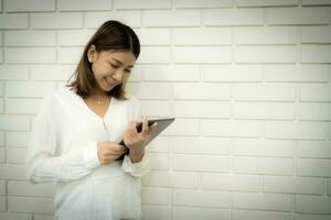 Beautiful asian business woman is standing and holding her tablet to check company profits with smile on her face against a white brick wall background. photo