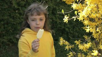 uma pequeno menina dentro amarelo é olhando às uma amarelo árvore e comendo gelo creme video