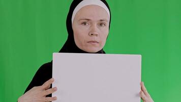 a woman in a black hijab stands in front of a green screen holding a plain board video