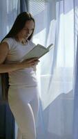 un embarazada mujer en blanco pantalones y un blanco camisa es en pie en frente de un ventana leyendo un libro video