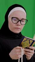 a woman wearing glasses and a black headscarf, holding a prayer beads and book. vertical video