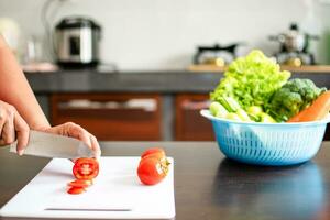 Close-up, A woman's hand was holding a knife and cutting tomatoes on a cutting board in the home kitchen. Cucumbers, lettuce, carrots in blue containers were placed on wooden table. Blurred background photo