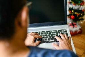 de cerca, personas utilizando ordenador portátil computadora dispositivos a hogar. un hombre mecanografía un teclado cuaderno computadora en el vivo habitación a hogar, blanco negro pantalla para diseño, texto, borroso fondo, y Navidad árbol foto