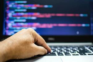 The programmer's hand is writing the source code. Programmers write programs for use with internet business services. The programming code displayed on the laptop screen. Closeup, Blurred background photo