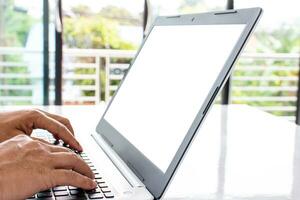 de cerca, personas trabajo hogar blogger un hombre se sienta y mecanografía un documento con un cuaderno computadora en un blanco mesa en el vivo habitación a hogar. el blanco pantalla es blanco para diseño, burlarse de arriba, recorte camino. foto