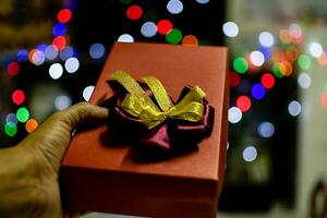 The hand of man holding red color Presents celebrates box with red and gold ribbons on Christmas day, take close-up photography, bokeh lights, and blurred background photo