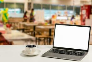 A blank white screen laptop and coffee cup are placed on a white desk in a food court. The Concept for business, technology, internet, design, and art. Closeup, selective focus, blurred background photo