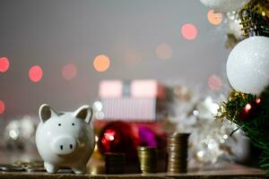 Closeup, White piggy banks, many coins on table wooden. Green Christmas tree decorated with medium balls. Concept investment budget wealth business during the festival. Selective focus on piggy banks photo