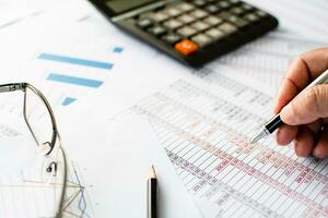 Financial accounting, work at home, Close-up Man's hand holding a pen examining accounting numbers. Numbers on paper and calculator, Many numbers, graph, pencil, glasses on paper, Blurred background. photo