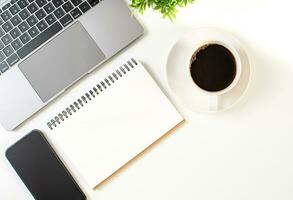 A coffee cup, a blank notebook, and a laptop are on a white table in the office. Working concept using technology, internet, computer. Copy space, Close up, top view, white background photo
