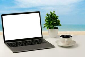A blank white screen laptop, coffee cup, and vase are placed on a white desk outdoor. concept for business, technology, internet, design, and programmer. Closeup, selective focus, sea, blur background photo