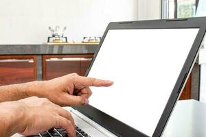 Closeup, A man's hand is a touch on a laptop blank screen to search out how to cook. notebook computer white screen on the table. Has a pot on the gas stove. Blurred background, moke up, clipping path photo