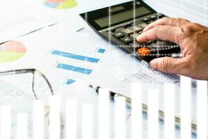 Man's finger pressing a calculator button To calculate financial transaction numbers, A pen, glasses on paper, Graphs showing statistics of financial, Financial accounting, Closeup, Blurred background photo