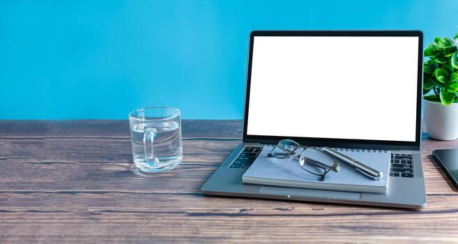 Blank, white screen laptop, drinking glass on a wooden table in the office. Working concept using technology, notebook, internet, laptop computer, network. Copy space on left, Closeup, blue background photo