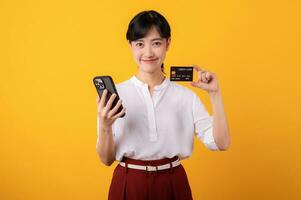 Portrait beautiful young asian woman enterpriser happy smile wearing white shirt and red plants holding credit card and smartphone isolation on yellow background. Online shopping concept. photo