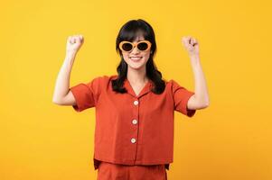 Portrait young beautiful asian woman happy smile dressed in orange clothes showing fist up hand gesture isolated on yellow studio background. Successful hooray celebrate young person concept. photo