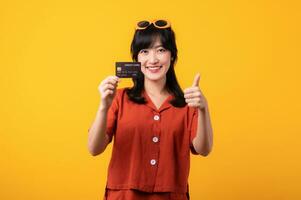 Portrait young beautiful asian woman happy smile dressed in orange clothes showing credit card and thumb finger up gesture isolated on yellow background. Pay and purchase shopping payment concept. photo