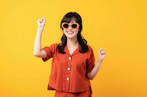 Portrait young beautiful asian woman happy smile dressed in orange clothes showing fist up hand gesture isolated on yellow studio background. Successful hooray celebrate young person concept. photo