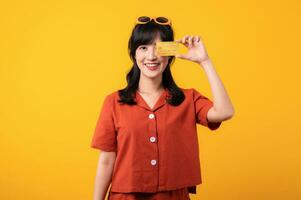 retrato joven hermosa asiático mujer contento sonrisa vestido en naranja ropa demostración crédito tarjeta aislado en amarillo antecedentes. pagar y compra compras pago concepto. foto