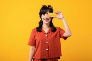 Portrait young beautiful asian woman happy smile dressed in orange clothes showing credit card isolated on yellow background. Pay and purchase shopping payment concept. photo