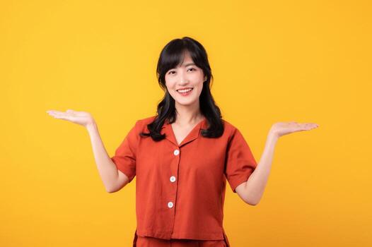 Portrait young beautiful asian woman happy smile dressed in orange clothes showing hand gesture, arm compare two variants demonstrate empty space isolated on yellow studio background. photo
