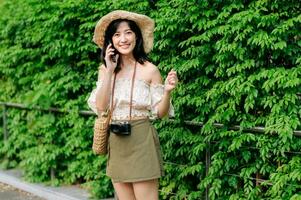 Portrait of asian young woman traveler with weaving hat, basket, mobile phone and camera on green public park background. Journey trip lifestyle, world travel explorer or Asia summer tourism concept. photo