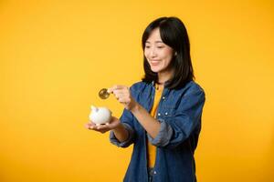 Happy young asian woman wearing yellow t-shirt denim shirt saving digital crypto currency to piggy bank isolated on yellow background. Digital money saving investment concept. photo