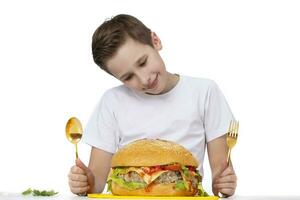 joven chico con grande hamburguesa aislado en blanco. un adolescente sostiene un dorado tenedor y un cuchara. foto