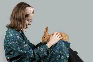 Beautiful little girl in glasses plays with a red rabbit on a gray background. photo