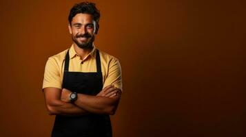 Portrait of a handsome indian barista standing with arms crossed against brown background. photo