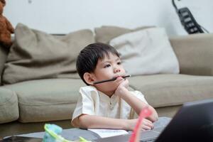 asiático chico sostiene lápiz en boca y muestra aburrido expresión mientras estudiando en línea en ordenador portátil foto
