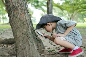 Asian boy wearing a hat in a forest exploration suit Use a magnifying glass to survey the tree area. photo