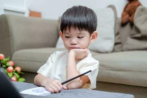Asian boy holding pencil and showing bored face while studying online on laptop photo