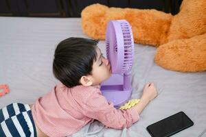 asiático chico acostado en el colchón en un caliente día jugando con un portátil ventilador felizmente foto