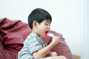 un asiático chico es comiendo delicioso hielo crema en un silla. foto