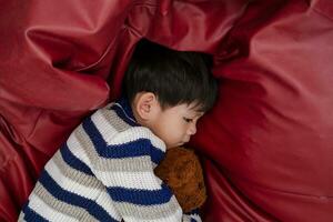 un chico es dormido y abrazando un osito de peluche oso en un rojo colchón. foto