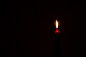 A single burning candle flame or light glowing on an orange candle on black or dark background on table in church for Christmas, funeral or memorial service with copy space photo