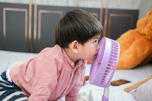 asiático chico acostado en el colchón en un caliente día jugando con un portátil ventilador felizmente foto