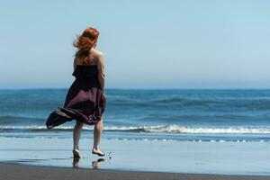 Rear view of woman in long dress walking on sandy beach towards water on background of blue sea photo