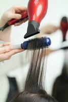 Hands of beautician drying brunette hair of client using hairbrush and hair dryer in beauty salon photo