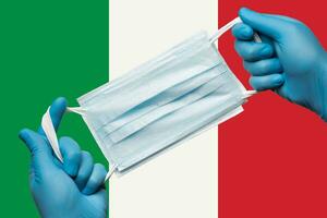 Medic holding respiratory face mask in hands in blue gloves on background flag of Italy or Italian Tricolour. Concept coronavirus quarantine, pandemic outbreak photo