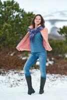 Young woman stands in winter forest near coniferous tree, looking at camera. Brunette woman with long hair, dressed in beige jacket, blue jeans, high-necked boots photo
